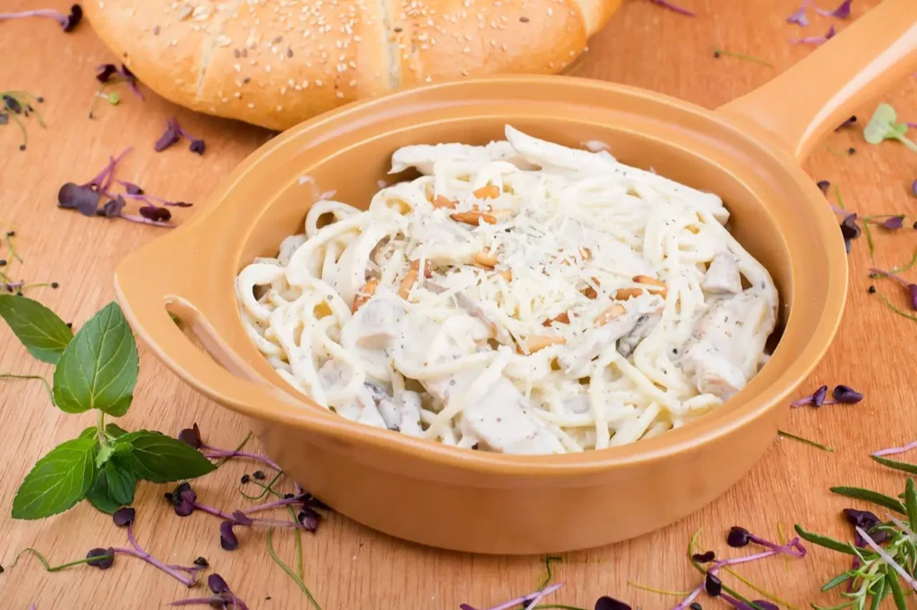 30-Minute-Chicken-Alfredo-Creamy. Creamy chicken Alfredo fettuccine pasta topped with grated cheese, served in a tan ceramic dish with a sesame bread roll and herbs on the side